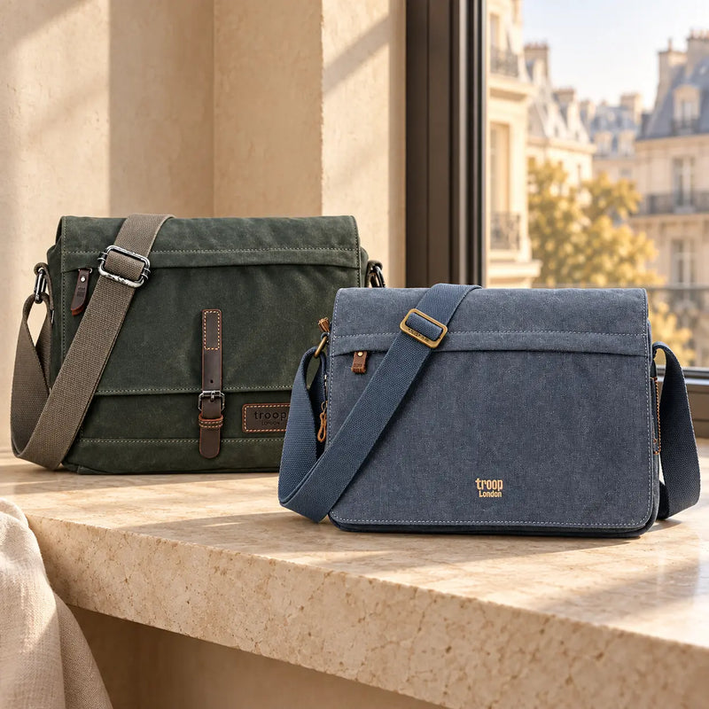 Green waxed canvas and blue canvas Troop London messenger bags on a pale stone bench beside a sunlit city window.
