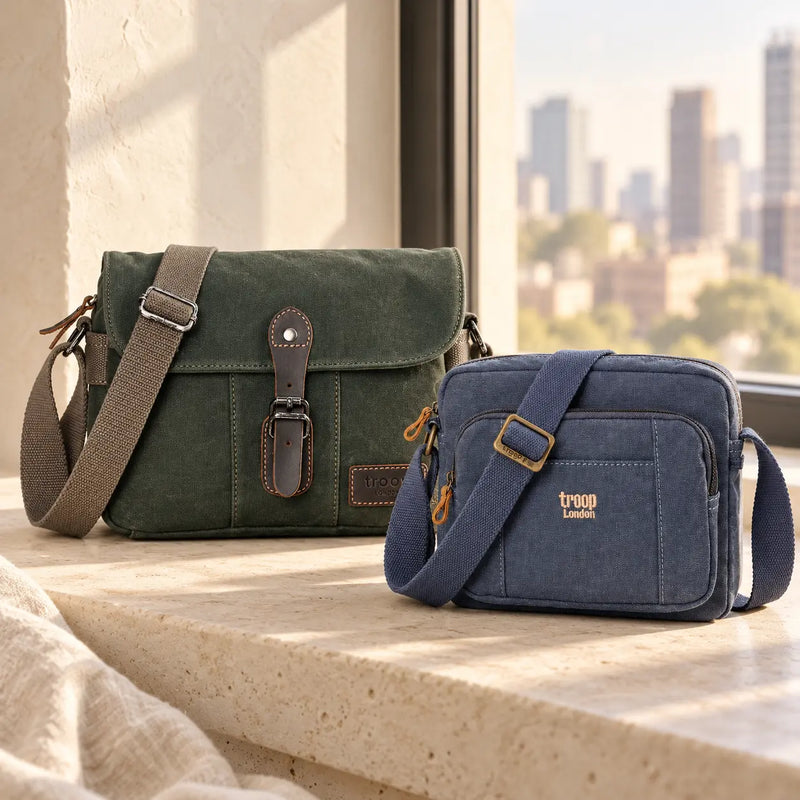 Green flap crossbody bag and compact blue zipped Troop London bag on a pale stone bench beside a sunlit window.