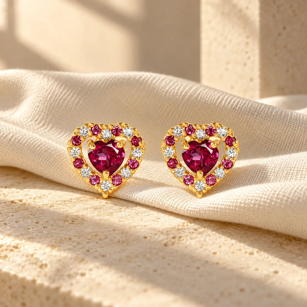 Rhodolite garnet heart stud earrings in gold vermeil resting on ivory linen in soft sunlight.