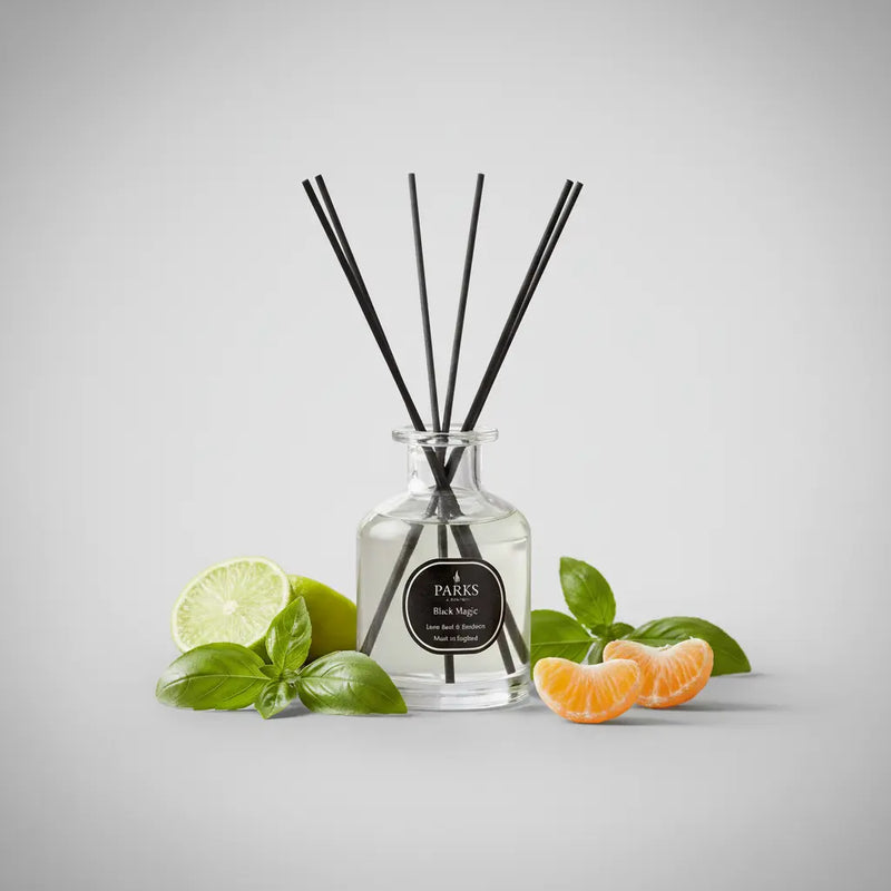 Parks London reed diffuser in clear glass apothecary bottle with black label and five black reeds, styled with fresh lime, basil leaves and mandarin segments on a silver backdrop