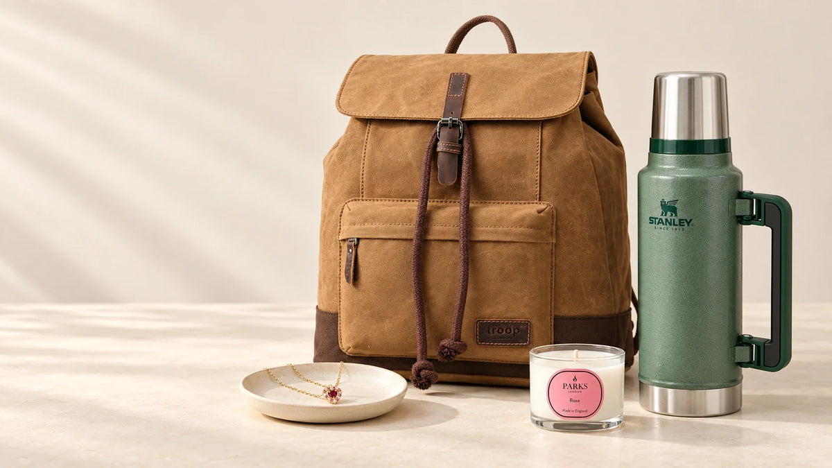 Troop London camel backpack, green Stanley bottle, Parks Rose candle and Vianne garnet heart necklace arranged on a warm ivory surface.