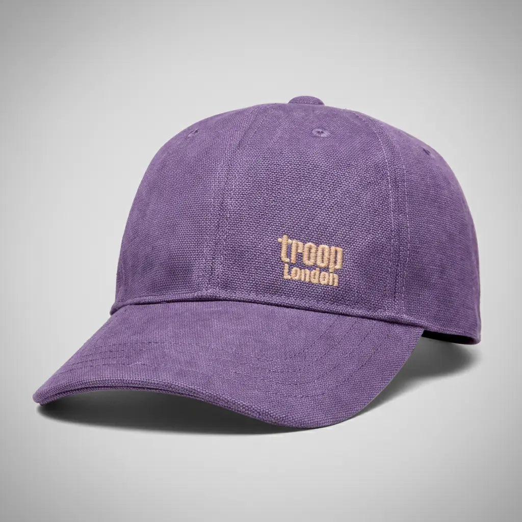 Troop London stone-washed canvas baseball cap in purple with gold embroidered logo, angled three-quarter view on a silver backdrop