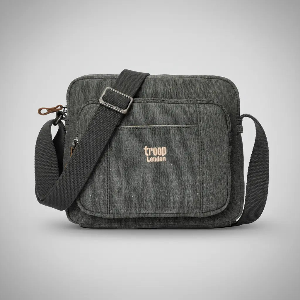 Troop London stone-washed canvas crossbody bag in charcoal with adjustable black strap and gold embroidered logo, front view on a silver backdrop