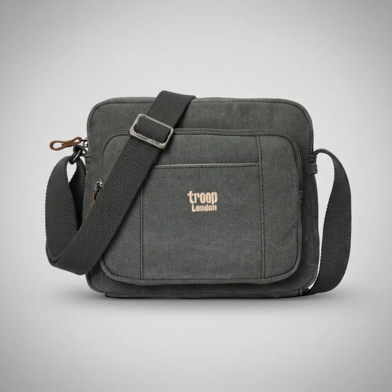 Troop London stone-washed canvas crossbody bag in charcoal with adjustable black strap and gold embroidered logo, front view on a silver backdrop