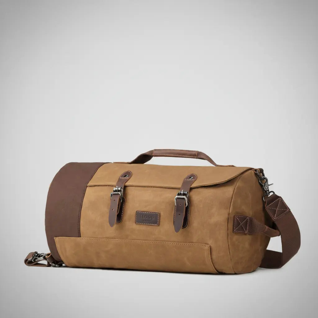 Troop London heritage canvas holdall bag in camel with dark brown leather trims, double buckle straps and detachable shoulder strap, angled view on a silver backdrop