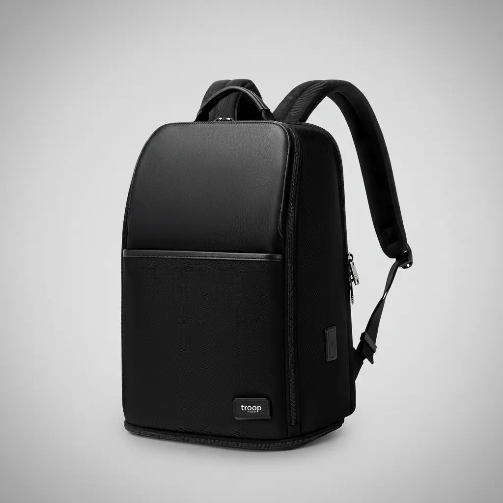 Troop London all-black business laptop backpack with leather-look top panel, front zip pocket and padded shoulder straps, angled view on a silver backdrop