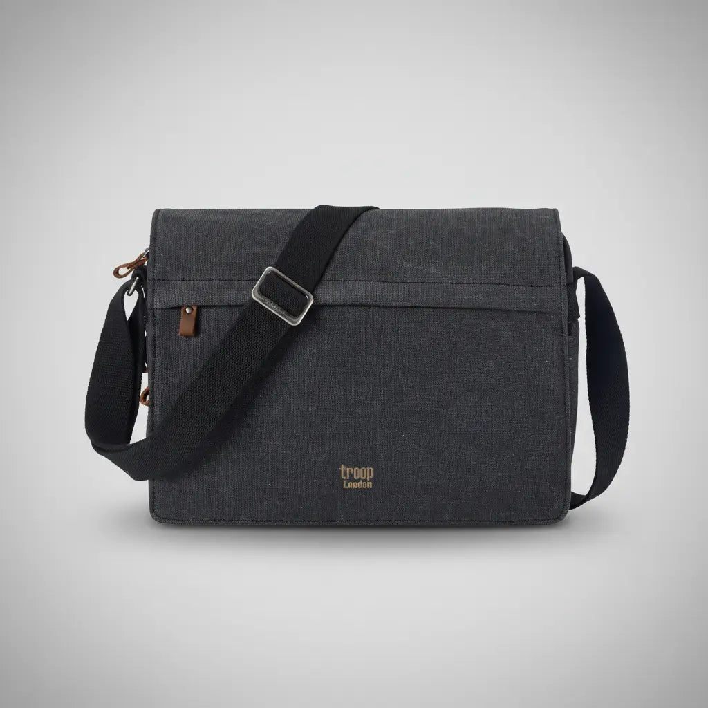 Troop London classic canvas messenger bag in charcoal with flap-over front, antique bronze zip pulls, tan leather tab and adjustable shoulder strap, front view on a silver backdrop