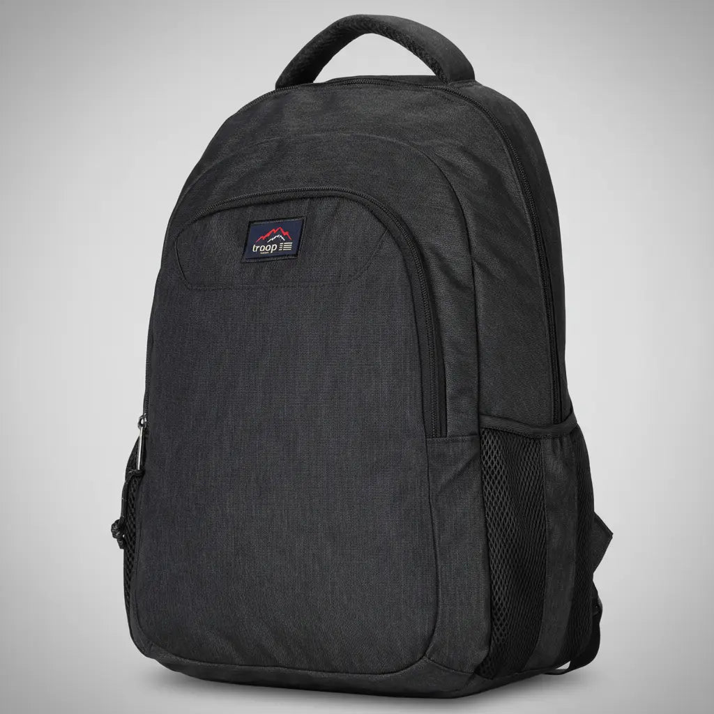 Troop London all-black urban daypack backpack with multiple zip compartments, mesh side pocket and padded shoulder straps, angled view on a silver backdrop
