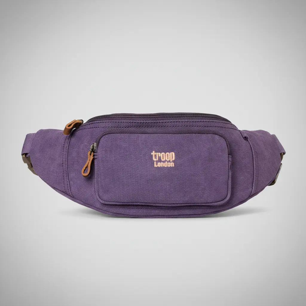 Troop London stone-washed canvas waist bag in purple with tan leather zip pull, front zip pocket and adjustable belt strap, front view on a silver backdrop
