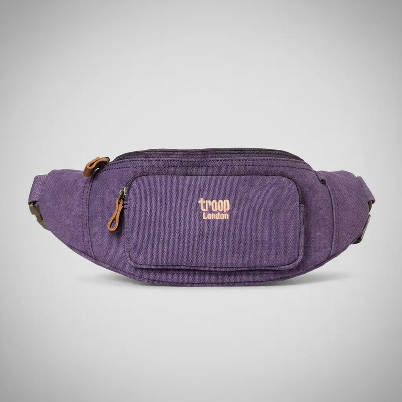Troop London stone-washed canvas waist bag in purple with tan leather zip pull, front zip pocket and adjustable belt strap, front view on a silver backdrop