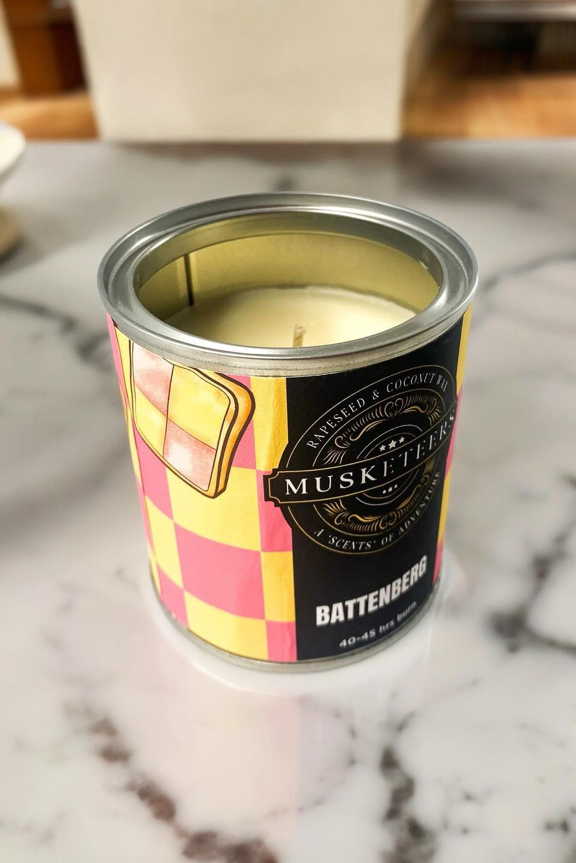 Musketeers Battenberg Scented Candle UK with sweet almond cake fragrance, showing a lit candle in a decorative container.