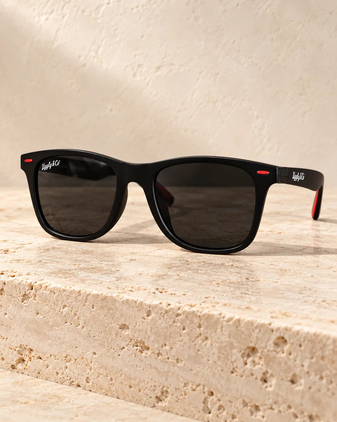 Black U&C Wayfarer sunglasses with dark lenses and red accents resting on pale travertine.
