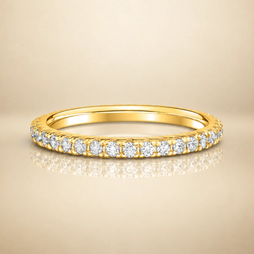A yellow gold eternity ring set with round brilliant-cut diamonds, shown on a warm beige reflective surface.