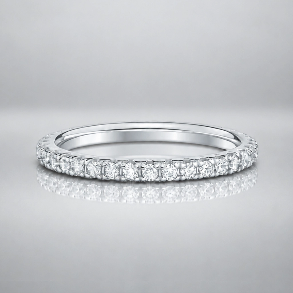 A thin silver eternity ring set with round brilliant-cut diamonds, shown on a reflective gray surface against a neutral gray background.