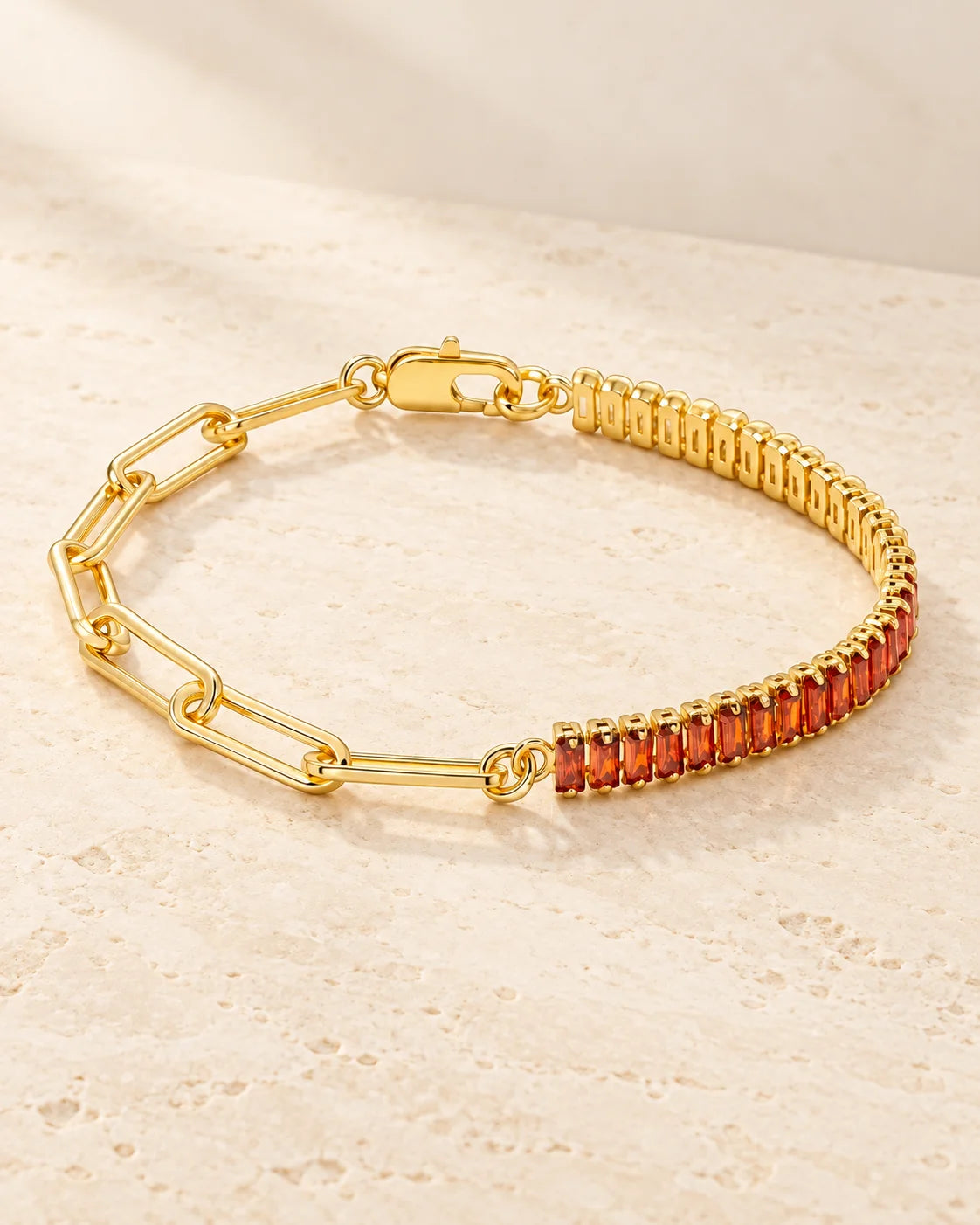 Gold plated bracelet with paperclip links and orange-red baguette stones arranged on pale travertine.