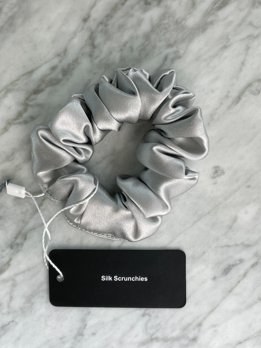 Silver silk scrunchie from DREAMSILK, showcasing a smooth, shiny fabric with a gentle, rounded shape on a plain background.