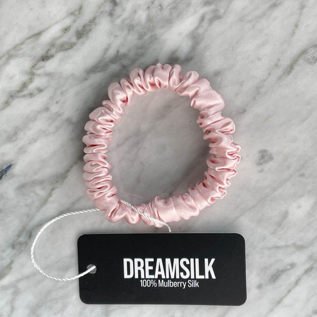 Pink silk scrunchie from DREAMSILK, skinny style, showing smooth, shiny silk fabric and elastic band on a neutral background.