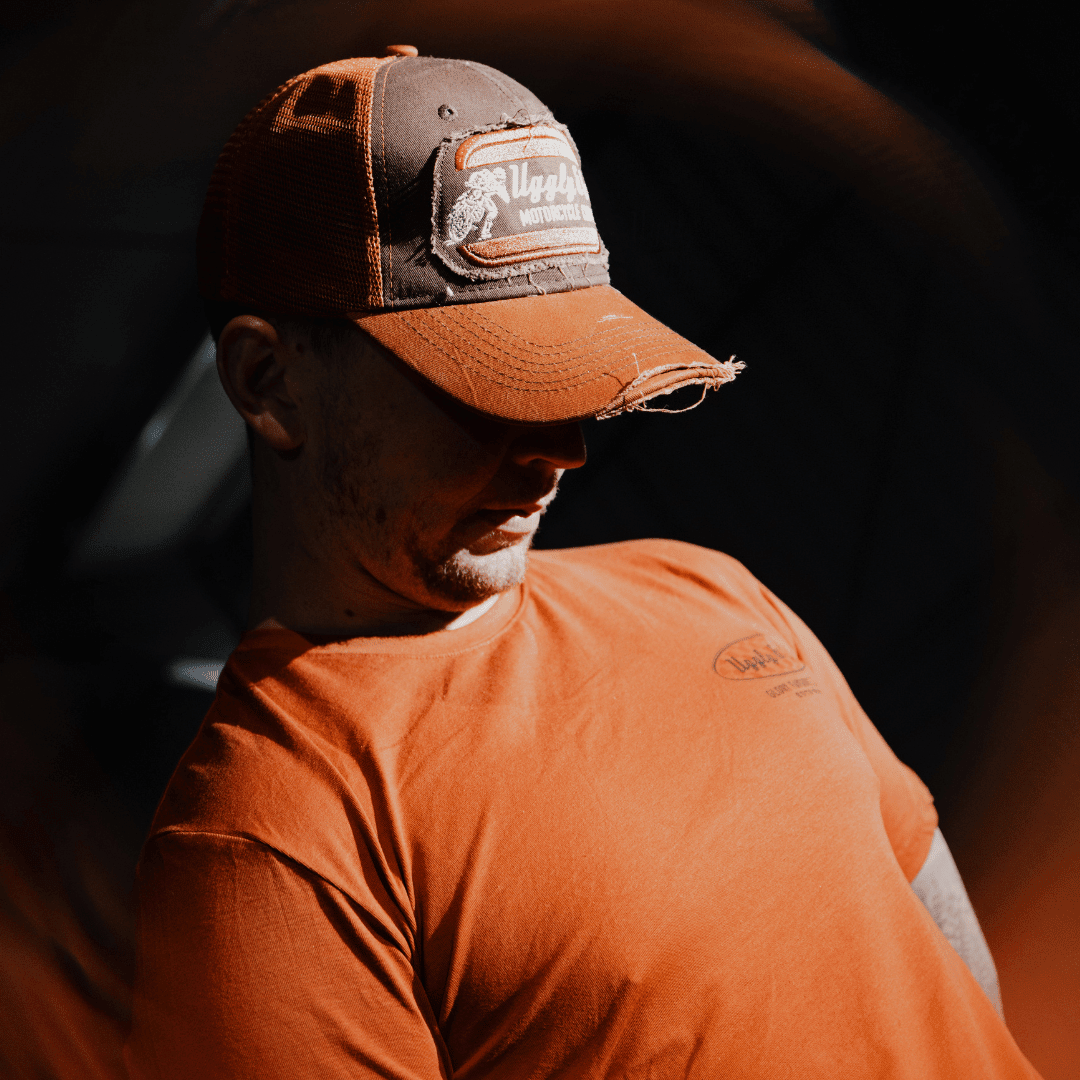 Rust-colored Uggly and Co flat track trucker cap with a retro biker design on the front, featuring mesh sides and adjustable snapback.