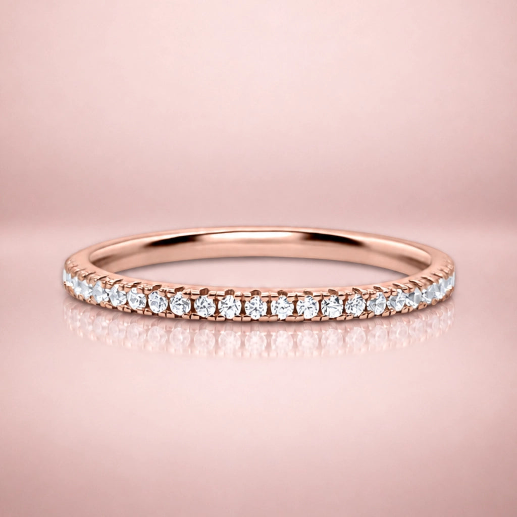 A rose gold eternity ring set with a row of round-cut clear gemstones, displayed on a soft pink reflective surface.