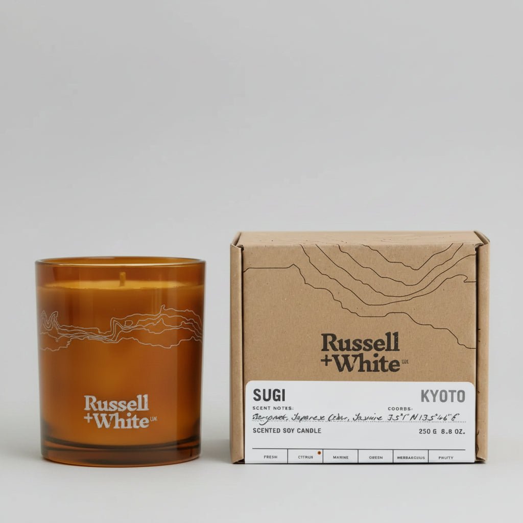 Russell + White Sugi scented soy candle in 250g jar with a natural wooden wick, surrounded by greenery and subtle lighting.