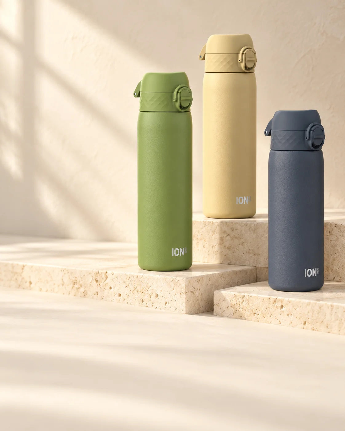 Khaki green, desert beige and ash navy ION8 water bottles arranged on pale stone ledges in soft sunlight.