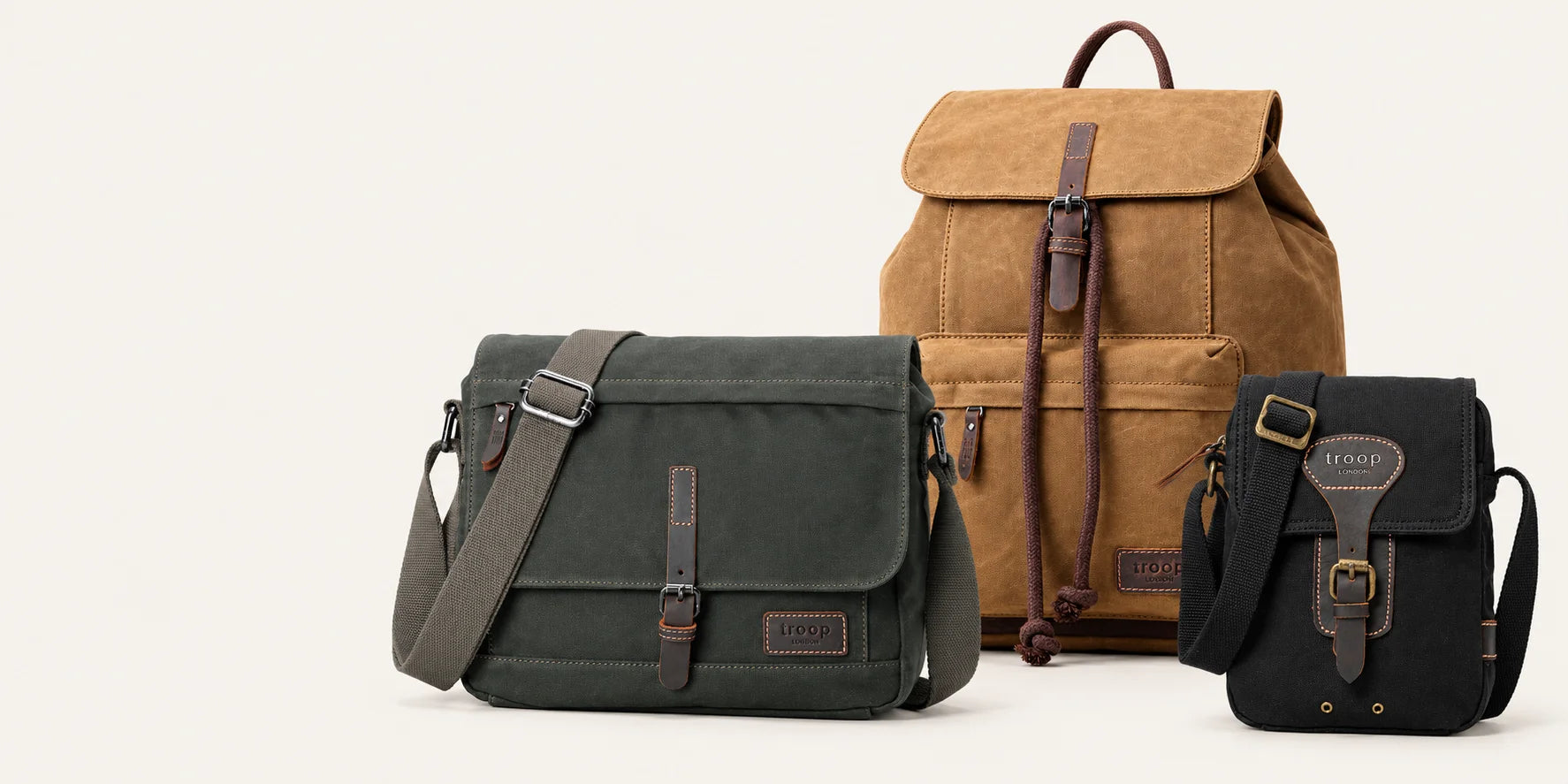 Three Troop London Heritage canvas bags: a camel backpack, dark-green messenger and black mini crossbody on an ivory background.