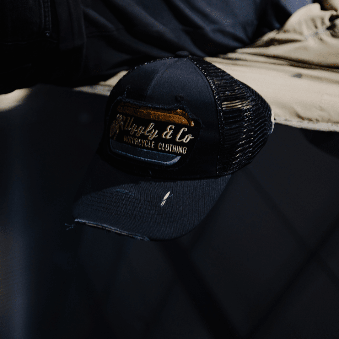 Black flat track trucker cap by Uggly and Co with a classic mesh back and curved brim, displayed on a motorcycle-themed background.