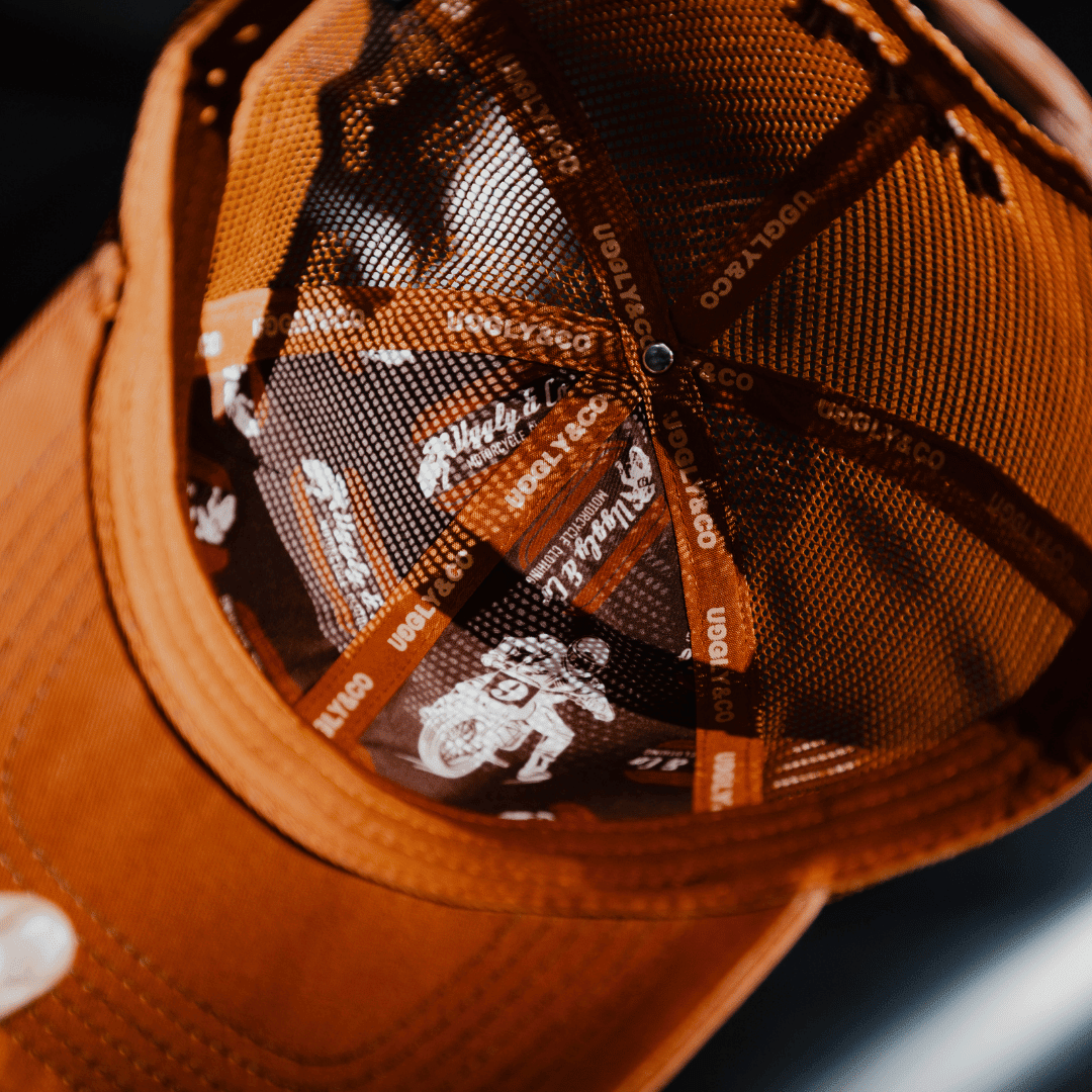 Rust-colored Uggly and Co flat track trucker hat with mesh back and curved brim, motorcycle-inspired design.