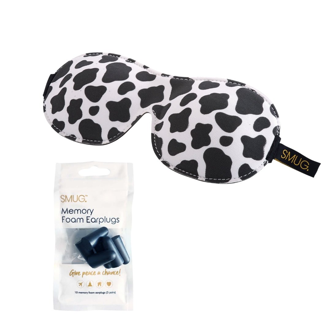 Contoured SMUG sleep mask with cow print design, black blackout fabric, and earplugs for complete sleep comfort.