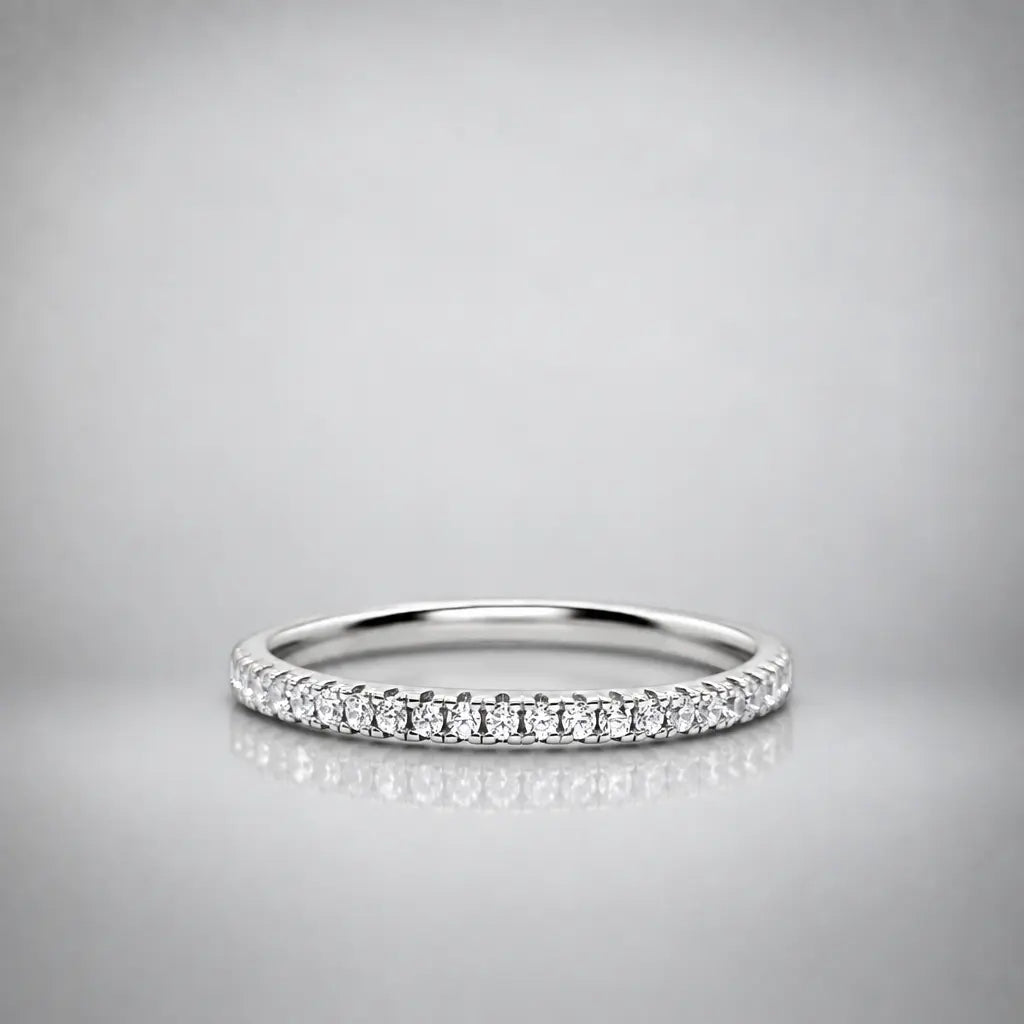 A thin silver diamond eternity ring with a row of round-cut diamonds set along the band, photographed on a neutral gray reflective surface.